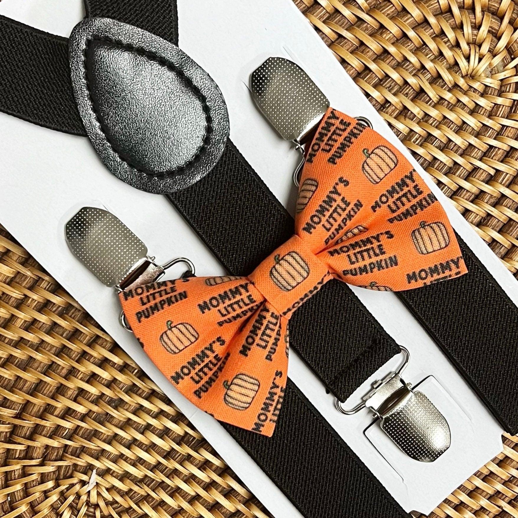 Orange pumpkin print bow tie with black suspenders on a white card against a woven mat background.