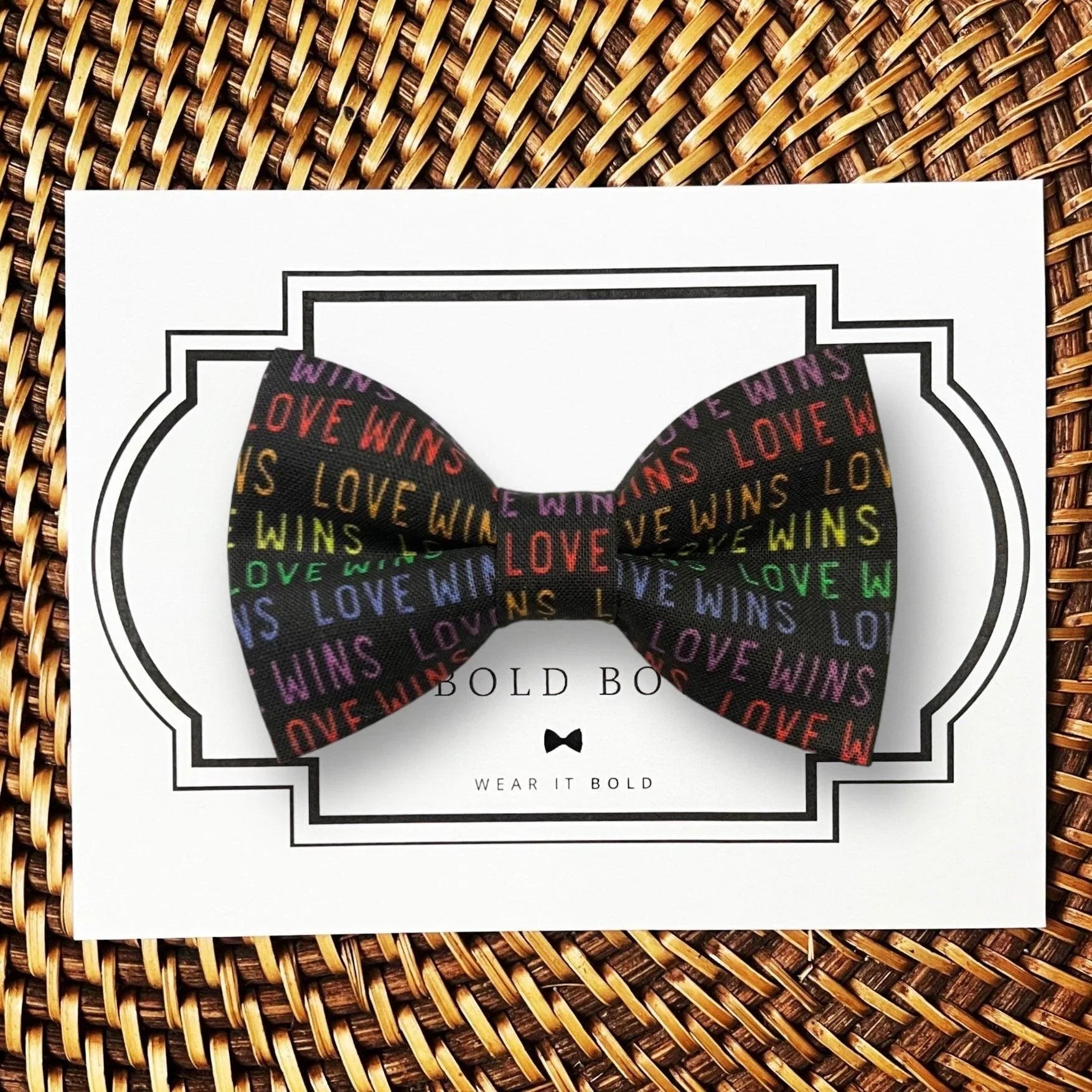 Love Wins Pride Bow for dog or cat collar displayed on a white card with a decorative frame against a woven background.