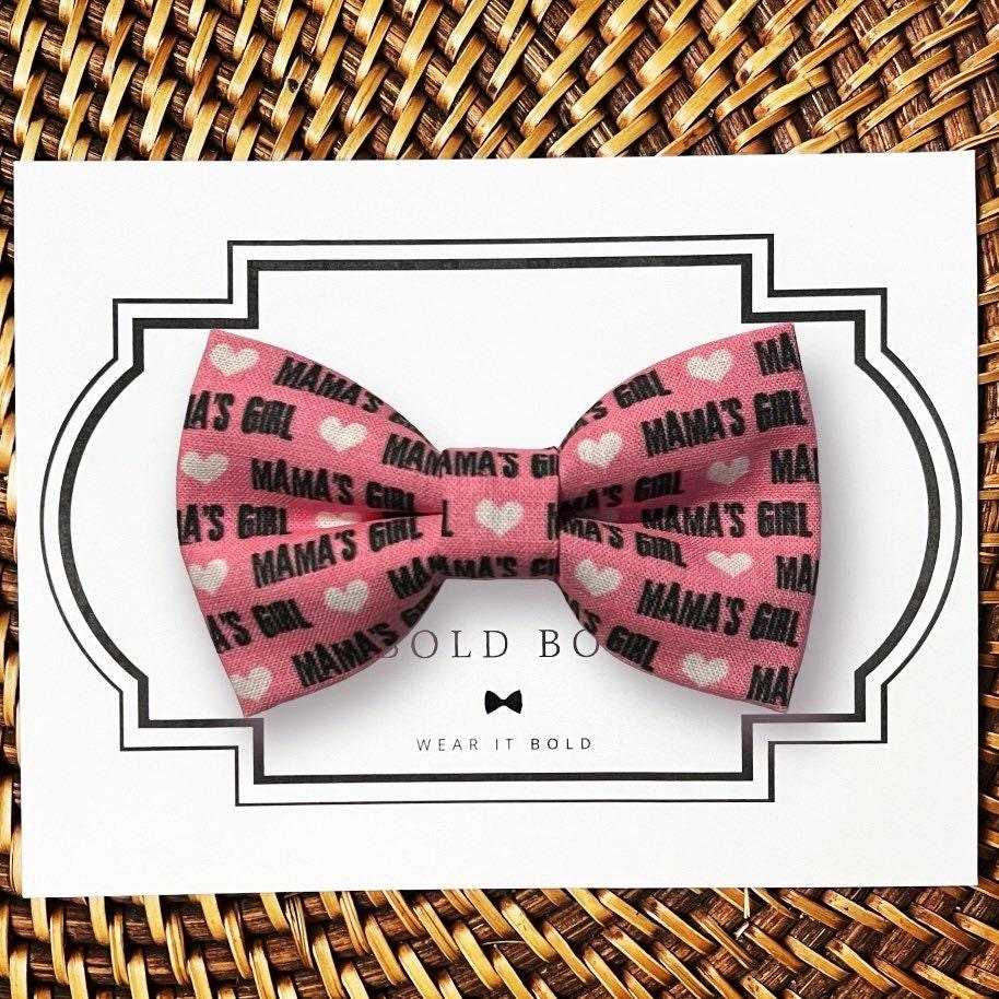 Pink dog bow tie or cat bow tie with 'Mama's Girl' text on a white card with a black border, placed on a woven surface.