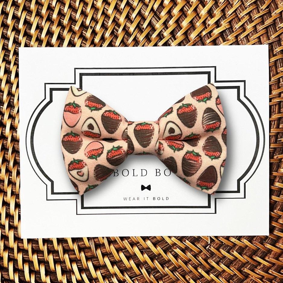 Pet Bow tie for Valentine’s Day with chocolate and strawberry pattern on a white card with 'The Bold Bow Tie’ branding, placed on a woven surface.