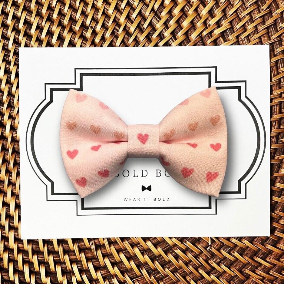 Valentine’s Day Pink bow tie with heart patterns on a white card with 'The Bold Bow Tie' branding, placed on a woven surface.