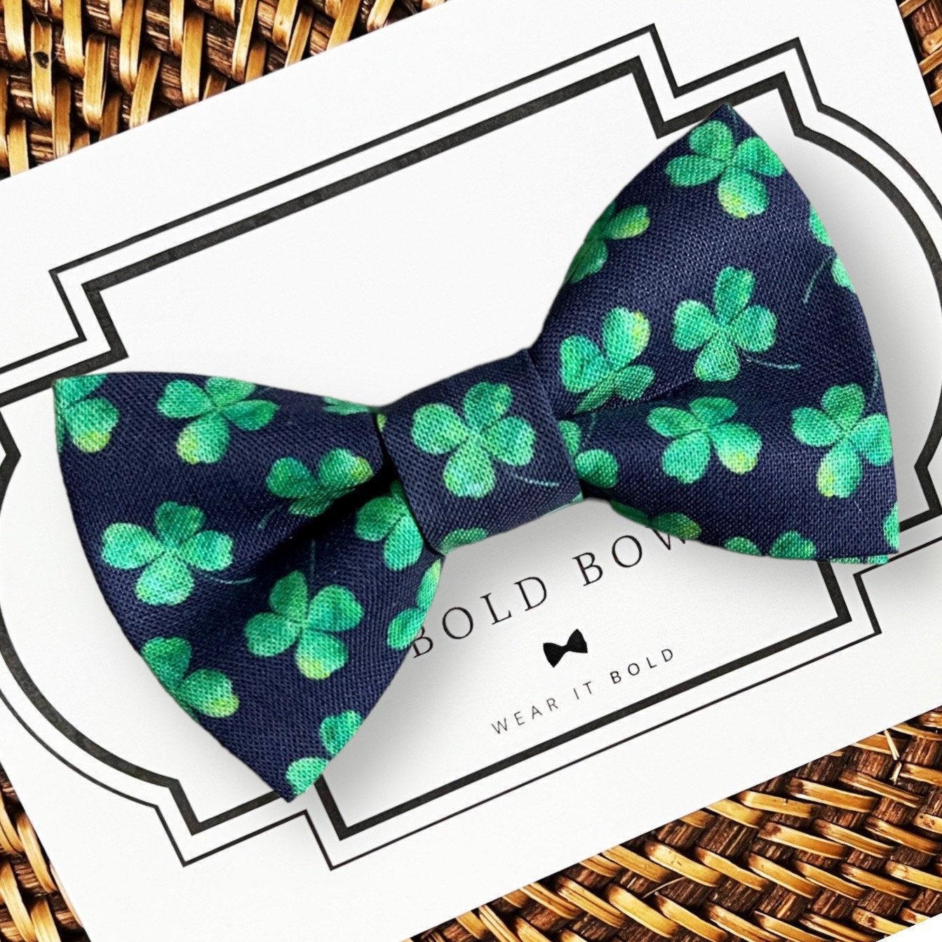 Navy blue dog bow tie for St. Patrick’s Day with green clover pattern on a white card with brand name, against a woven background.