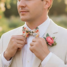 A man in a white suit and floral bow tie is adjusting his bow tie, with a pink rose boutonniere pinned to his lapel.