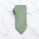 A sage green tie for men’s wedding necktie with a matching fabric neck loop, laid flat on a textured white surface.
