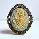 May flower Lily of the Valley brooch with blue and ivory floral design and ornate frame on a white background