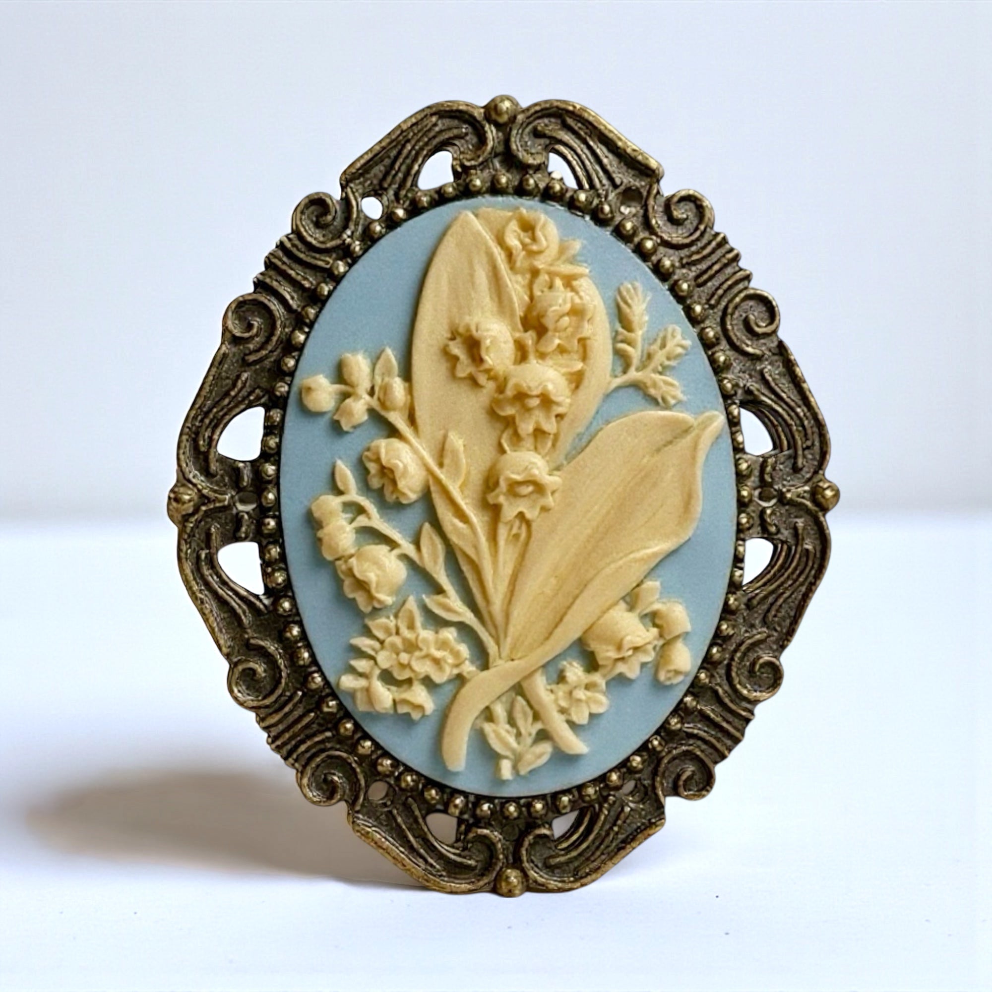 May flower Lily of the Valley brooch with blue and ivory floral design and ornate frame on a white background