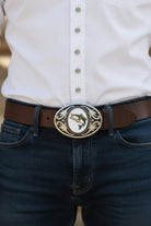 Person wearing a fish belt buckle, white shirt, dark jeans, and a brown belt.
