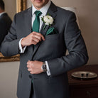A groomsman in an emerald green tie is adjusting his pocket square, with a white flower boutonniere pinned to his lapel.