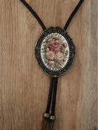 A decorative floral pendant necklace with a black leather tassel hanging from it, displayed on a wooden surface.