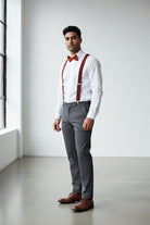 Man wearing a white shirt, gray pants, burnt orange bow tie, terracotta suspenders, and brown shoes as a groomsman in a wedding.