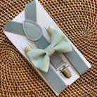Sage green bow tie and grey suspenders set on a white box with gold clasp, placed on a woven surface.