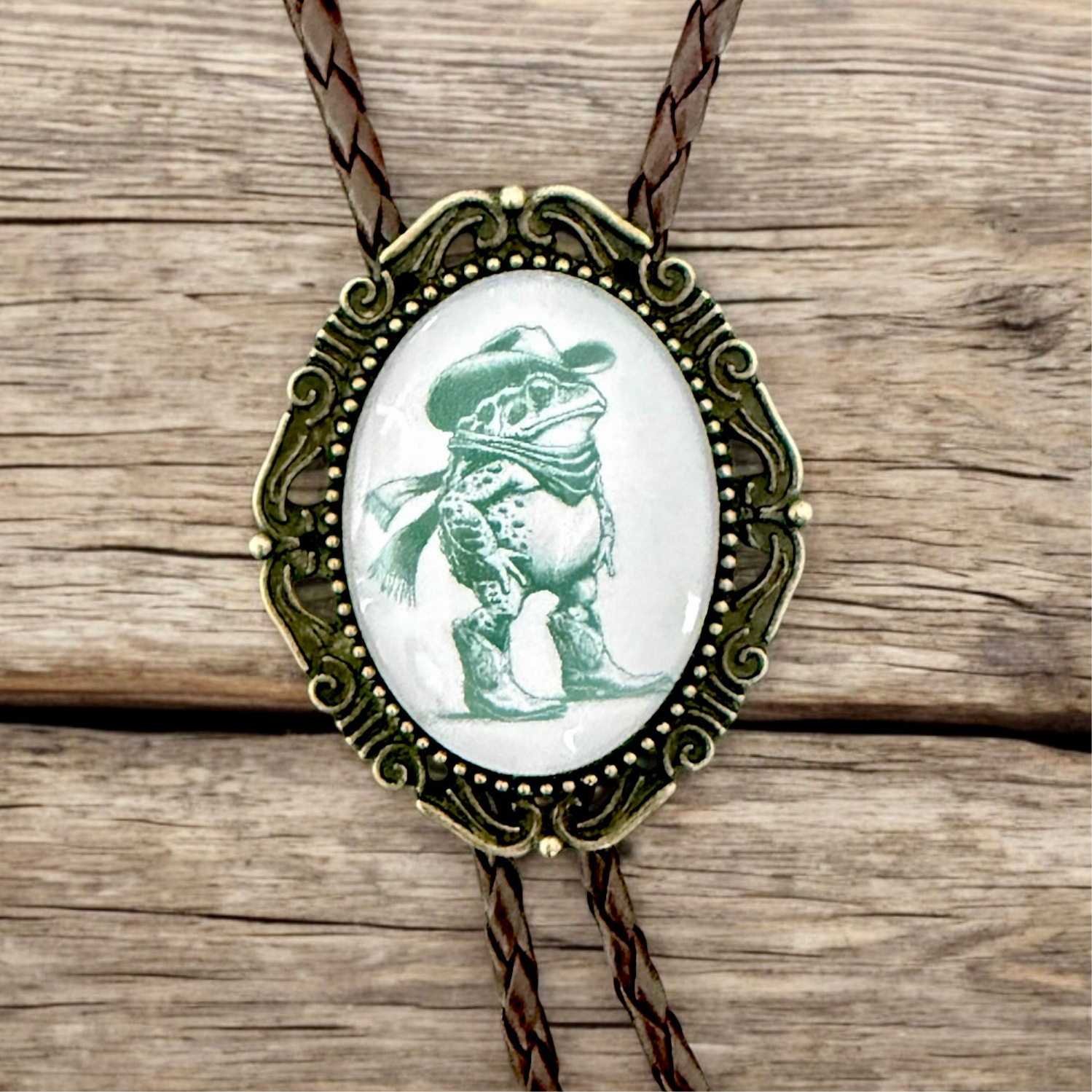 Bolo tie with a frog wearing a cowboy hat on an oval pendant, set against a wooden background.