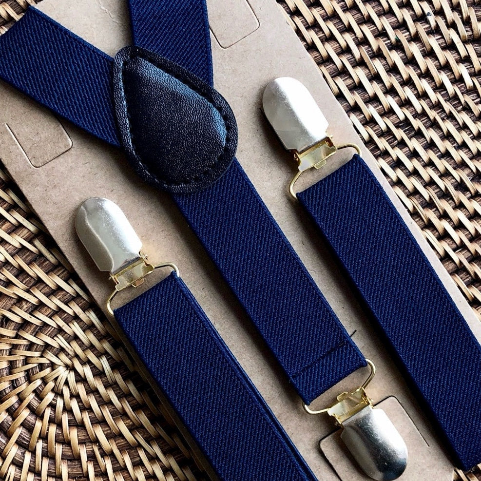 Set of navy suspenders with silver clips for groomsmen, ring bearer outfit, and wedding attire on a woven surface.