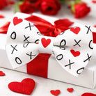 White Valentines Day bow tie with red hearts and black 'XO' pattern on a red and white gift box with roses in the background.