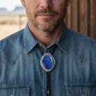 Man wearing a blue denim shirt with a decorative bolo tie featuring a blue stone.