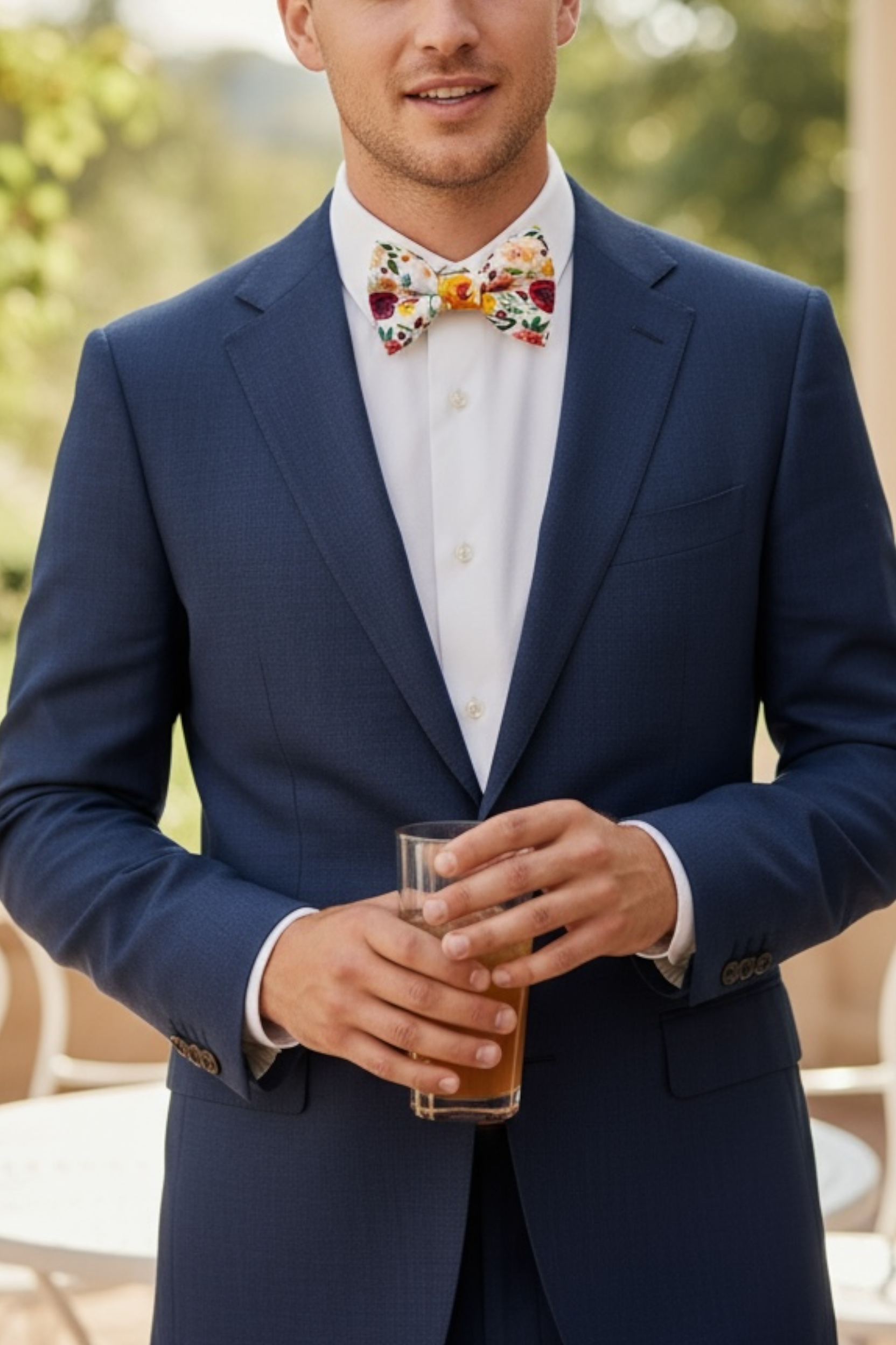 Groomsman in a navy suit with a colorful bow tie holding a glass outdoors.