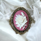 Vintage-style brooch with white rose design on a pink background, set against a white lace fabric.