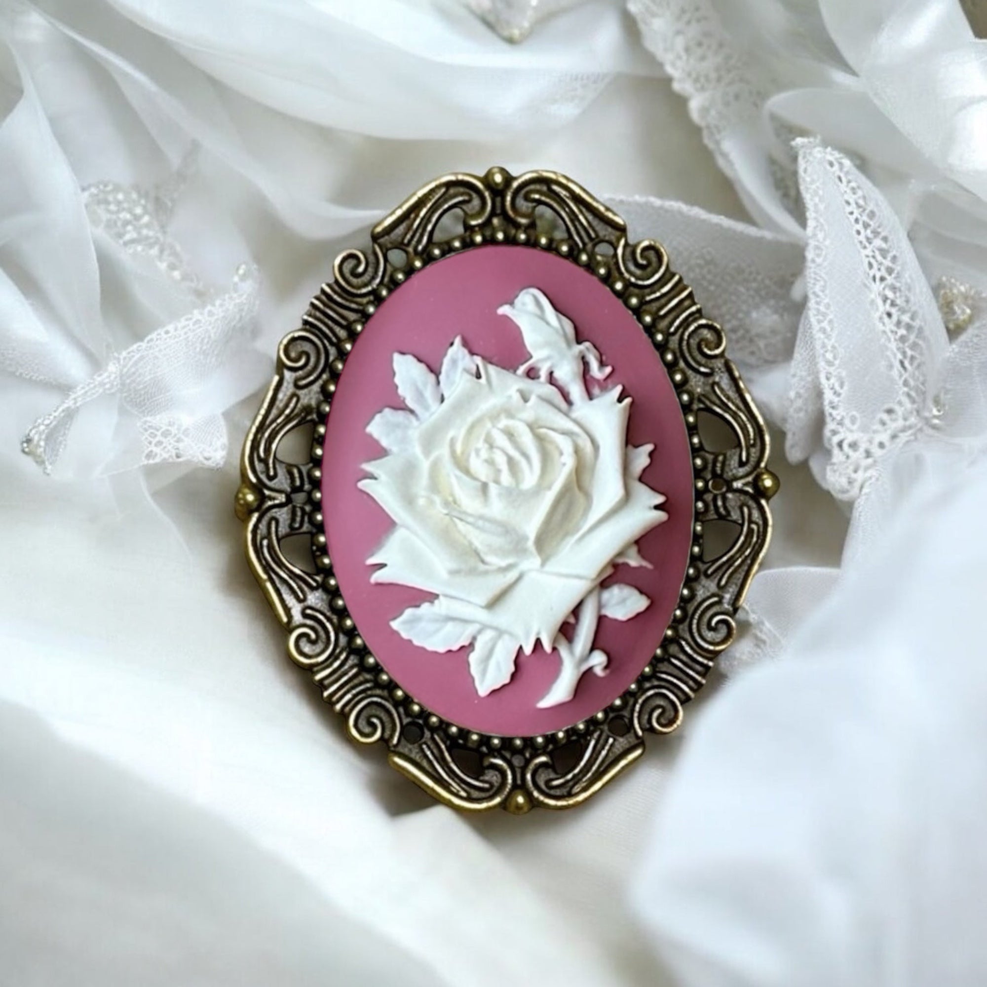 Vintage-style brooch with white rose design on a pink background, set against a white lace fabric.