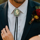 Man wearing a dark suit with a white shirt, decorative May flower green lily of the valley bolo tie, and floral boutonniere.