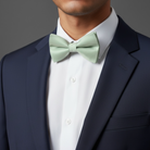 Man wearing a navy suit with a white shirt and wedding Sage green bow tie on a gray background