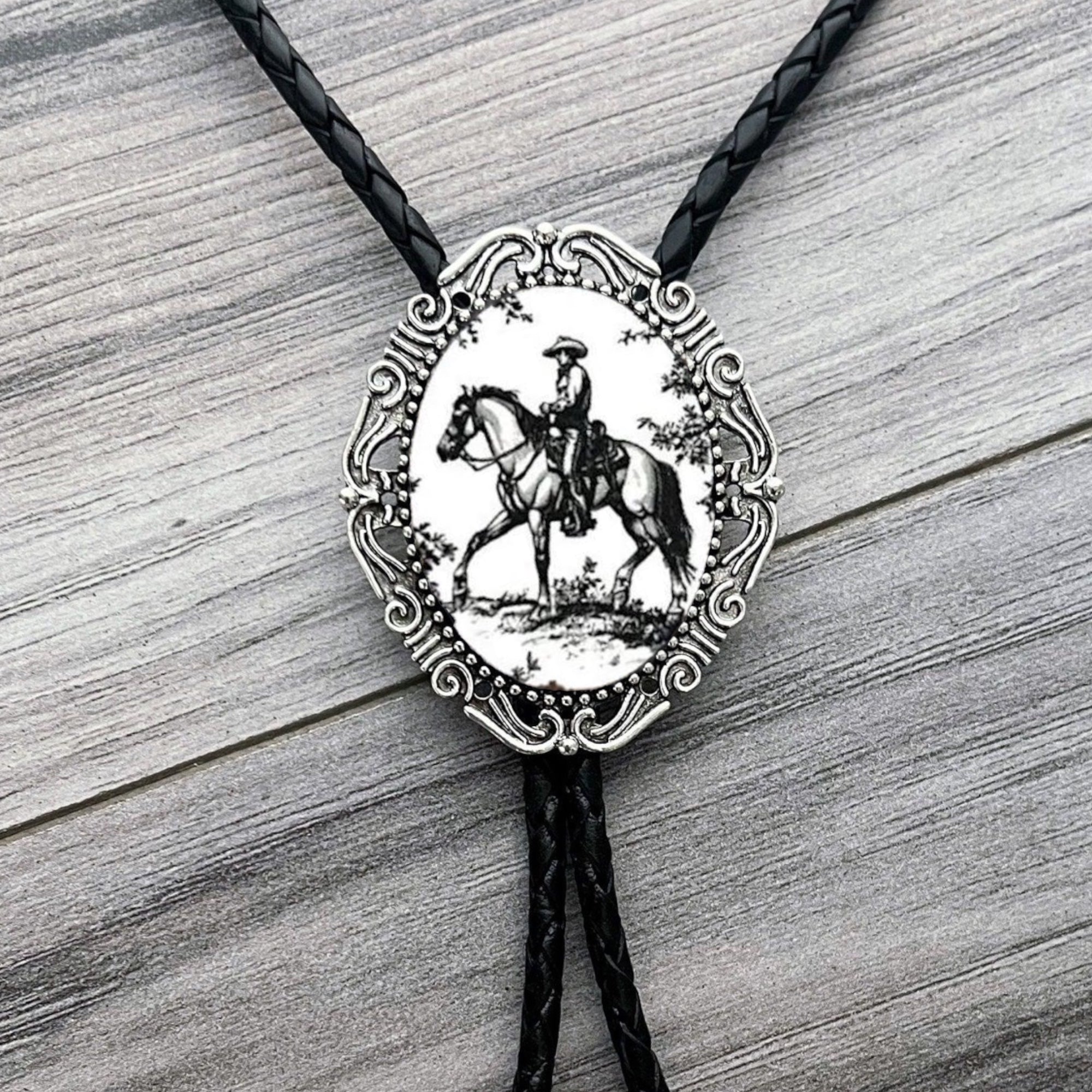 Black and white Cowboy Bolo tie with a decorative pendant featuring a horse and rider on a wooden surface