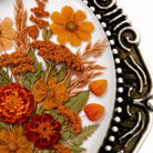 a close-up view of a decorative plate with a variety of orange and red flowers, along with dried flower petals and leaves. The plate has an ornate, decorative metal frame that adds an elegant touch to the overall presentation.