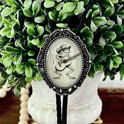 A western-style bolo tie with a black cord and a silver-toned oval pendant featuring a frog playing guitar, displayed in front of green foliage.