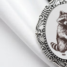 a close-up view of a decorative metal frame with a raccoon illustration on it, placed on a white fabric surface.
