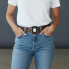 Person wearing blue jeans with a decorative flower belt buckle against a neutral background