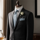 A mannequin wearing a black suit jacket, light blue tie, and a small white flower in a light blue boutonniere.