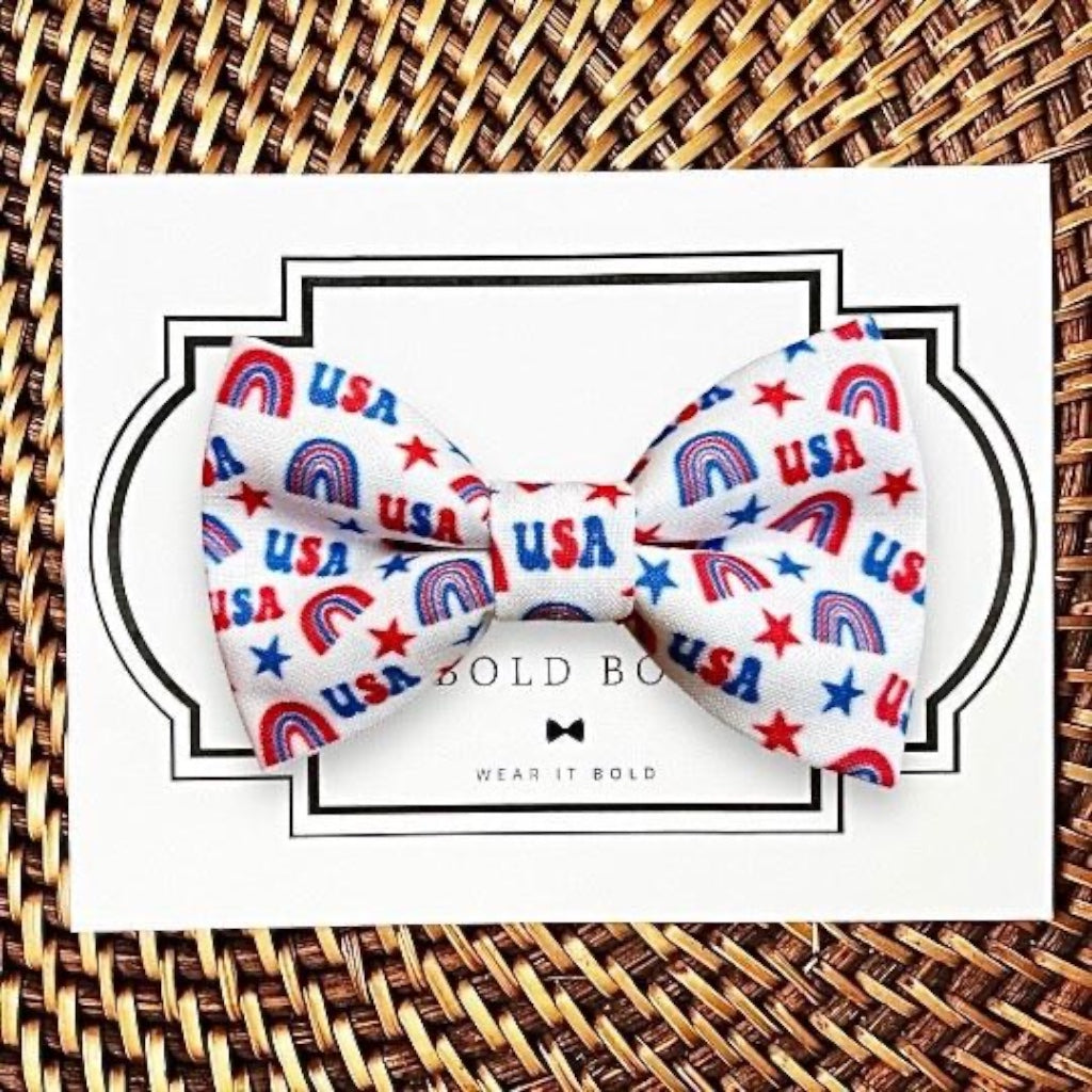 Dog Bow tie with 'USA' and rainbow pattern on a white card with 'The Bold Bow Tie' branding, placed on a woven surface.