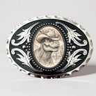 Close-up of a silver and black oval Western belt buckle featuring a T-Rex dinosaur wearing a cowboy hat in a desert landscape.