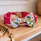 A pink collar with a floral print dog bow tie and a gold leash are displayed on a wooden surface.