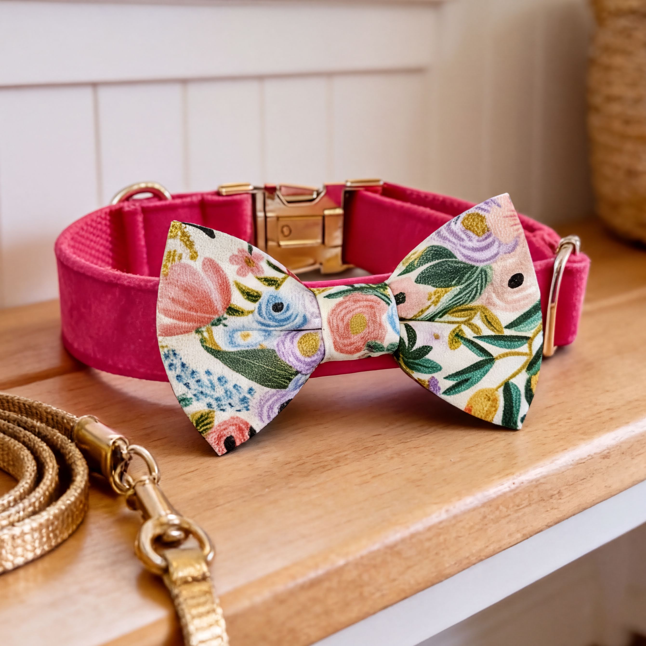 A pink collar with a floral print dog bow tie and a gold leash are displayed on a wooden surface.