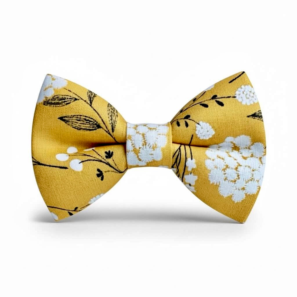 A pre-tied mustard floral bow tie in yellow fabric with a white floral and black leaf pattern, shown against a plain white background.