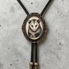 A close-up of a decorative pendant featuring an image of an opossum, with a black cord and gold-colored tassels.