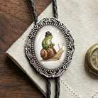 A close-up of a bolo tie necklace with a black braided cord and an ornate silver oval pendant. The pendant features an illustration of a green frog sitting on a brown snail wearing a small black hat, set against a light background.