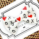 White Valentine’s Day dog bow tie with red hearts and black 'XO' text on a card with 'Wear It Bold' branding.