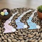 Three decorative Easter bunny tags with names on a woven mat with Easter eggs and a nest.