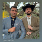Two well-dressed men posing for a photo in a wooded area, one wearing a blue plaid suit and the other wearing a brown suit and a black cowboy hat.