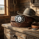 A pair of brown leather belts and a gray cowboy hat are displayed on a wooden dresser, with a decorative metal buckle featuring a cow design.