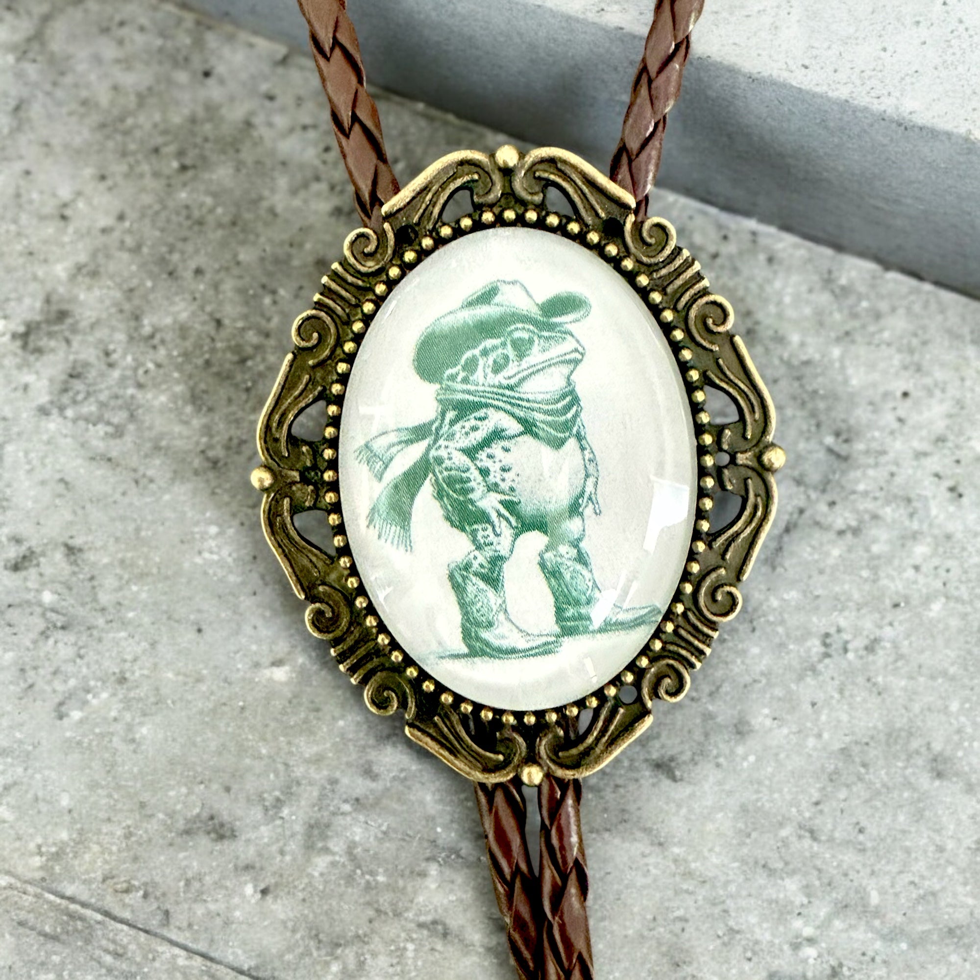 Bolo tie with decorative pendant featuring a frog in a cowboy hat on a concrete surface