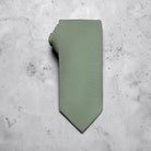 A sage green tie for men’s wedding necktie with a matching fabric neck loop, laid flat on a textured light gray concrete surface.