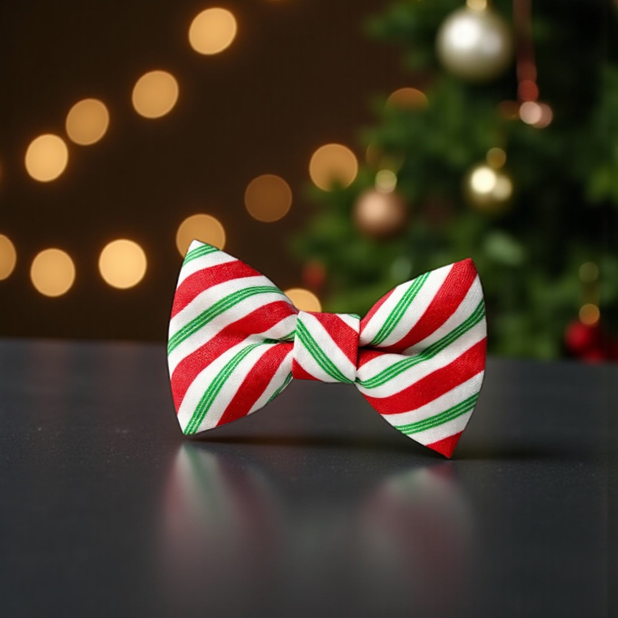 Red, green, and white striped bow tie on a dark surface with a blurred Christmas tree and lights in the background.
