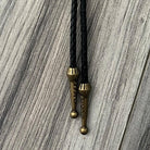 a close-up view of a black braided leather tie with brass-colored metal ends, hanging on a wooden surface.