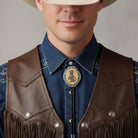 Man wearing a raccoon gun bolo tie and brown leather vest with fringe and a blue shirt against a gray background