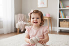 Toddler in a pink dress and pink hair bow with hearts holding a teddy bear in a room with a bookshelf and toys.