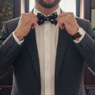 Man adjusting a black and gold bow tie wearing a suit with a blurred background