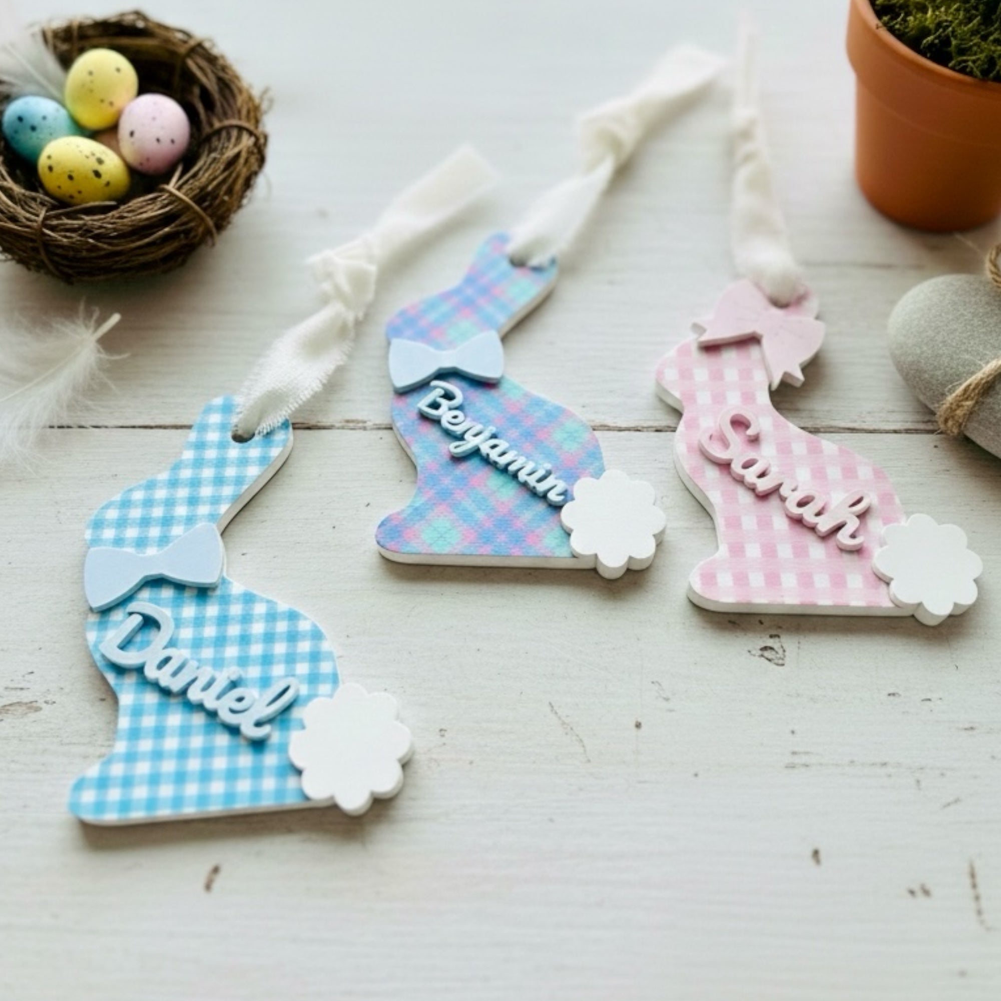 A collection of pastel personalized acrylic Easter tags in bunny shapes with 3D names, arranged on a light wood surface with Easter eggs