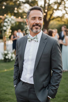 Groomsman in a gray suit with a dark green floral bow tie standing outdoors at a wedding.